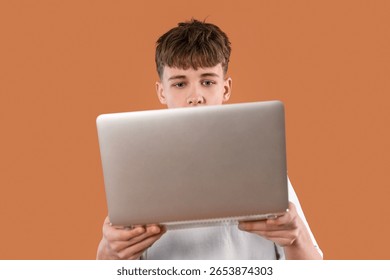 Young boy focused on using laptop in a casual indoor setting with a warm orange background during daytime