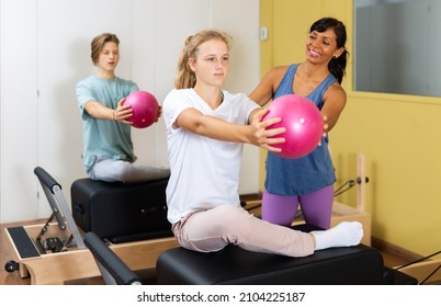 Young boy and girl doing exercises on pilates reformers and using small fitness balls. Their trainer hispanic woman correcting them.