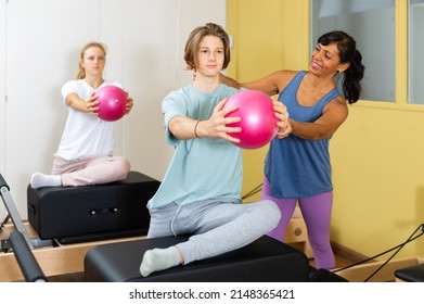 Young boy and girl doing exercises on pilates reformers and using small fitness balls. Their trainer hispanic woman correcting them.