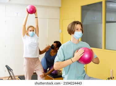 Young boy and girl in face masks doing exercises on pilates reformers and using small fitness balls. Their trainer hispanic woman correcting them.