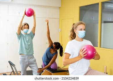Young boy and girl in face masks doing exercises on pilates reformers and using small fitness balls. Their trainer hispanic woman correcting them.