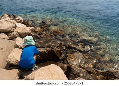 Young boy in green hat and blue swimsuit exploring rocky seashore by the clear blue water on a sunny summer day. Concept of childhood adventure, nature discovery, and outdoor activities by the sea.  - Powered by Shutterstock