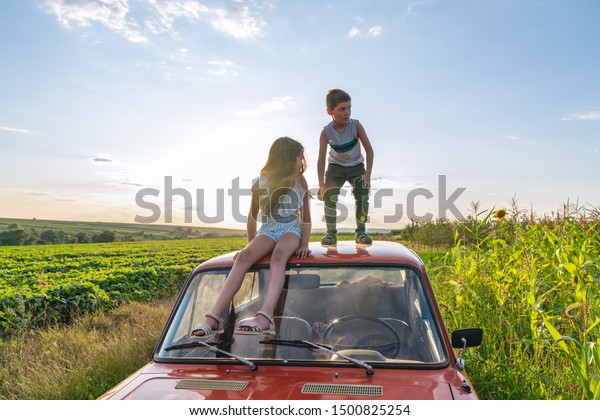Young boy in grey t-shirt standing on the roof of the\
red retro car and looking scary at the left side her sister in blue\
striped dress sitting beside and looking at him, field and blue sky\
on the