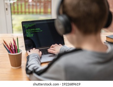 Young boy hacking and coding system on computer laptop screen with green program code text typing on keyboard selective focus on fingers wearing headphones sat at desk