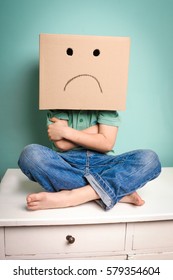 Young boy has a cardboard box over his head with sad emoticon drawn on it and is sitting on a white drawer cabinet