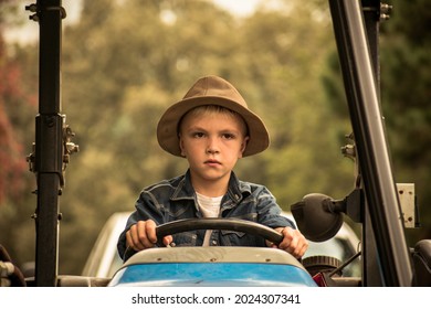 A Young Boy In A Hat Driving A Tractor- A Little Farm Boy Driving A Tractor- Helping Out On The Farm- A Young Child In A Vintage Hat Driving A Blue Tractor With Both Hands On The Steering Wheel