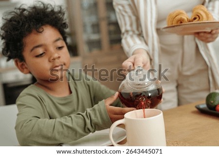 Young boy having breakfast while adult pouring tea into his cup in cozy kitchen environment. He appears enthusiastic and attentive, adding warmth to the morning scene