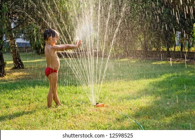 Young Boy Having Fun Playing With Water From A Sprinkler In The Garden