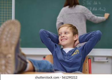 Young boy with his feet up on the desk and a mischievous grin sitting in the classroom as the teacher writes on the blackboard with her back to him - Powered by Shutterstock