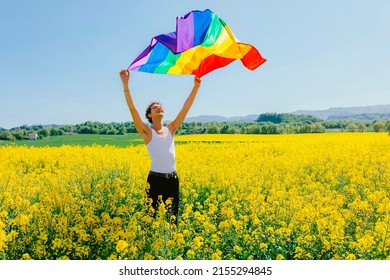 Young Boy Holding The LGBT Gay Rainbow Flag At Sunset, On Yellow Rapeseed Field In Spring. Happiness, Freedom And Love Concept For Same Sex Couples. Lifestyle Outdoors.