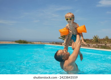 A young boy with inflatable orange arm bands and sunglasses is joyfully lifted up by an adult male in a sunlit outdoor swimming pool - Powered by Shutterstock