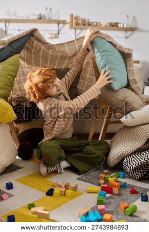 A young boy joyfully constructs a vibrant fort using cushions and blankets during a sunny afternoon.