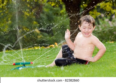 Young Boy Or Kid Cools Off By Playing In Water Sprinkler At Home In His Back Yard On Hot Summer Day