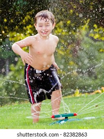 Young Boy Or Kid Cools Off By Playing In Water Sprinkler At Home In His Back Yard On Hot Summer Day