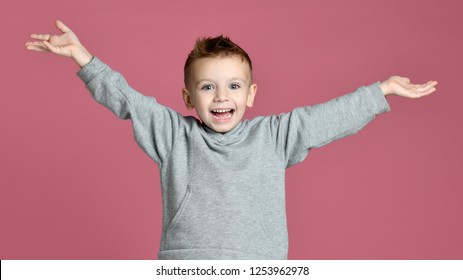 Young Boy Kid Jumping In Grey Hoodie With Hands Spread Up Laughing Smiling On Pink Background