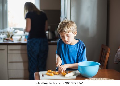 Young boy in the kitchen cutting fruits during prepering coctail, in the background his mother