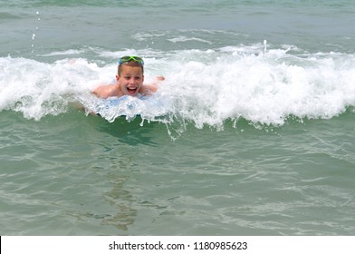 Young Boy Laughs As He Rides A Small Wave At Madeira Beach, Florida.  He Has Green Snorkle Glasses On His Head.