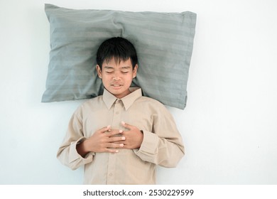 A young boy is laying on his stomach with his hands on his chest. He is wearing a shirt and a grey pillow. The boy appears to be in a state of distress or discomfort - Powered by Shutterstock