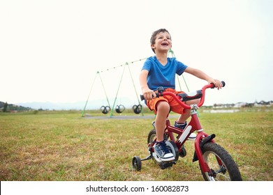 Young Boy Learning To Ride A Bike With Bicycle Training Wheels In Park