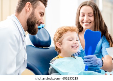 Young boy looking at the mirror with toothy smile sitting on the chair with dentist and assistant at the dental office
