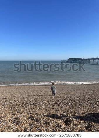 Young boy looks out to sea on a pebbled beach.  Beach peer in the background stretches out to see with a beautiful blue sky, very peaceful and tranquil 