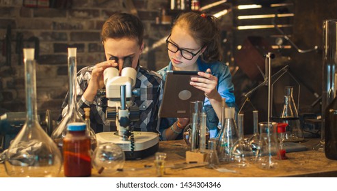 Young Boy Is Making Chemistry Experiments While Girl Is Looking At A Tablet Computer In A Garage At Home.