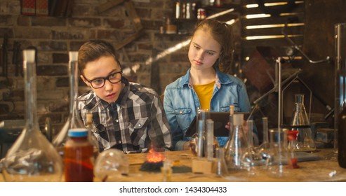 Young Boy Is Making Chemistry Experiments While Girl Is Holding A Tablet Computer In A Garage At Home.