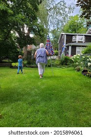 A Young Boy And An Older Woman Walking Alongside Each Other On The Lawn In A Suburban Backyard. The Lady Is Holding A Closed Beach Umbrella And The Child Is Holding A Long Stick.