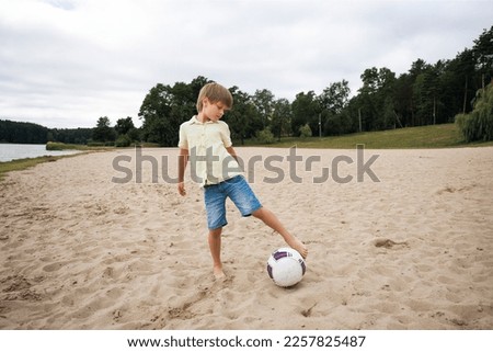 A young boy on the beach runs towards a soccer ball and wants to score a goal on a warm summer day. He is wearing denim shorts and a yellow shirt.