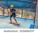 Young boy on a waterboard rides a wave pool indoors, wearing a bright yellow helmet and holding onto a red rope