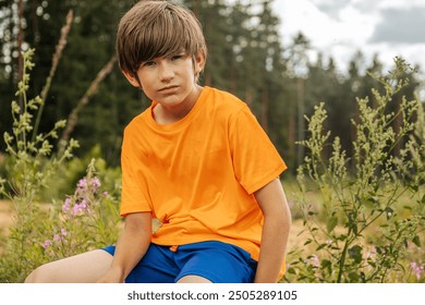 A young boy in an orange shirt and blue shorts sits outside with a serious expression on his face. The background boasts greenery and forest, emphasizing the natural, peaceful environment. - Powered by Shutterstock