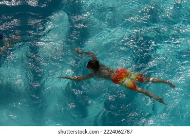 A Young Boy In Orange And Yellow Swim Trunks Floats With His Face Underwater In A Swimming Pool On A Sunny Day.