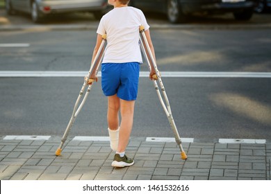 Young Boy In Orthopedic Cast On Crutches Walking On The Street Near The Road. Child With A Broken Leg On Crutches, Ankle Injury. Bone Fracture And Ankle Fracture In Children