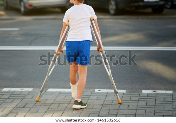 Young boy in\
orthopedic cast on crutches walking on the street near the road.\
Child with a broken leg on crutches, ankle injury. Bone fracture\
and ankle fracture in\
children