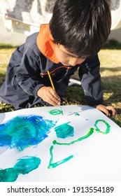 Young Boy Painting With A Brush On A White Surface With Blue Paint, Wearing A Smock, In The Yard On A Sunny Day With Clothes Lying In The Background