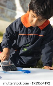 Young Boy Painting With A Brush On A White Surface With Blue Paint, Wearing A Smock, In The Yard On A Sunny Day With Clothes Lying In The Background