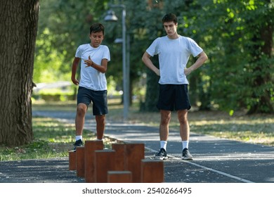 Young boy participates in outdoor physical activity, balancing on wooden stumps. Engaging in this fun exercise, he builds coordination and strength, supervised by a trainer in a natural setting. - Powered by Shutterstock