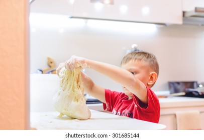 Young boy playing with dough while baking covering his arms in the uncooked mixture with a funny expression on his face as he stands at the kitchen counter