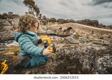 A young boy is playing with a toy truck on a rocky hillside. The boy is wearing a blue jacket and he is enjoying himself. The scene is peaceful and serene - Powered by Shutterstock