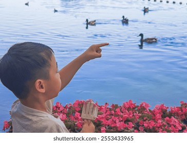 Young boy points at ducks in water, holding ice cream near vibrant pink flowers. - Powered by Shutterstock