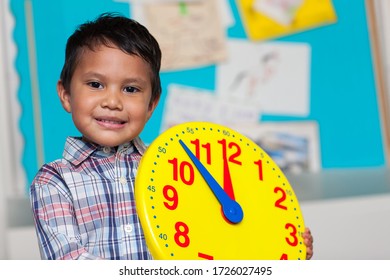 Young Boy With A Preppy Shirt, Holding A Big Analog Clock With A Colorful Bulleting Board In The Background.