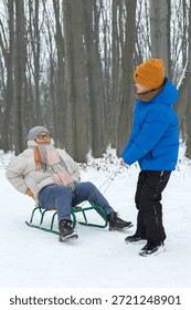 young boy pulls his elderly grandmother on a sled during a walk in a snowy park. intergenerational bonding, role reversal, active aging and family joy outdoors. Real life, authentic family connection 