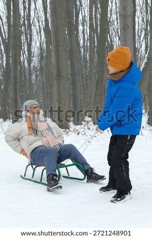 young boy pulls his elderly grandmother on a sled during a walk in a snowy park. intergenerational bonding, role reversal, active aging and family joy outdoors. Real life, authentic family connection 