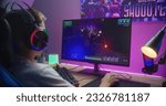 Young boy puts on headphones and starts play third person shooter. Gamer enjoys online video game on home personal computer. Desk illuminated by RGB LED strip light. Gaming at home concept. Back view.