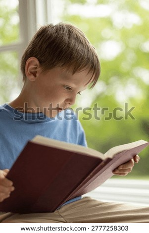 A young boy reading a book in front of a window