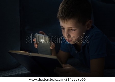 Young boy reading book at night with a lamp from mobile phone