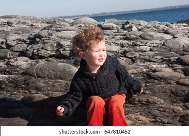 Young Boy Is Relaxing Against Rocks On A Beach In Newport, Rhode Island. He Is Dressed For Fall Or Spring Weather In A Black Sweater And Orange Pants.  He Has Wavy, Curly Hair. 