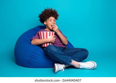 Young boy relaxing on a bean bag chair holding a popcorn bucket against a vibrant blue background