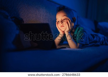 Young boy resting on sofa, experiencing digital eye strain and exhaustion while looking at a glowing tablet screen in a dark room