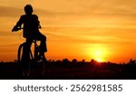 A young boy rides his bicycle down a rural road on his way back from school during the quiet of the late evening. The golden hues of the setting sun paint the landscape in warm colors.